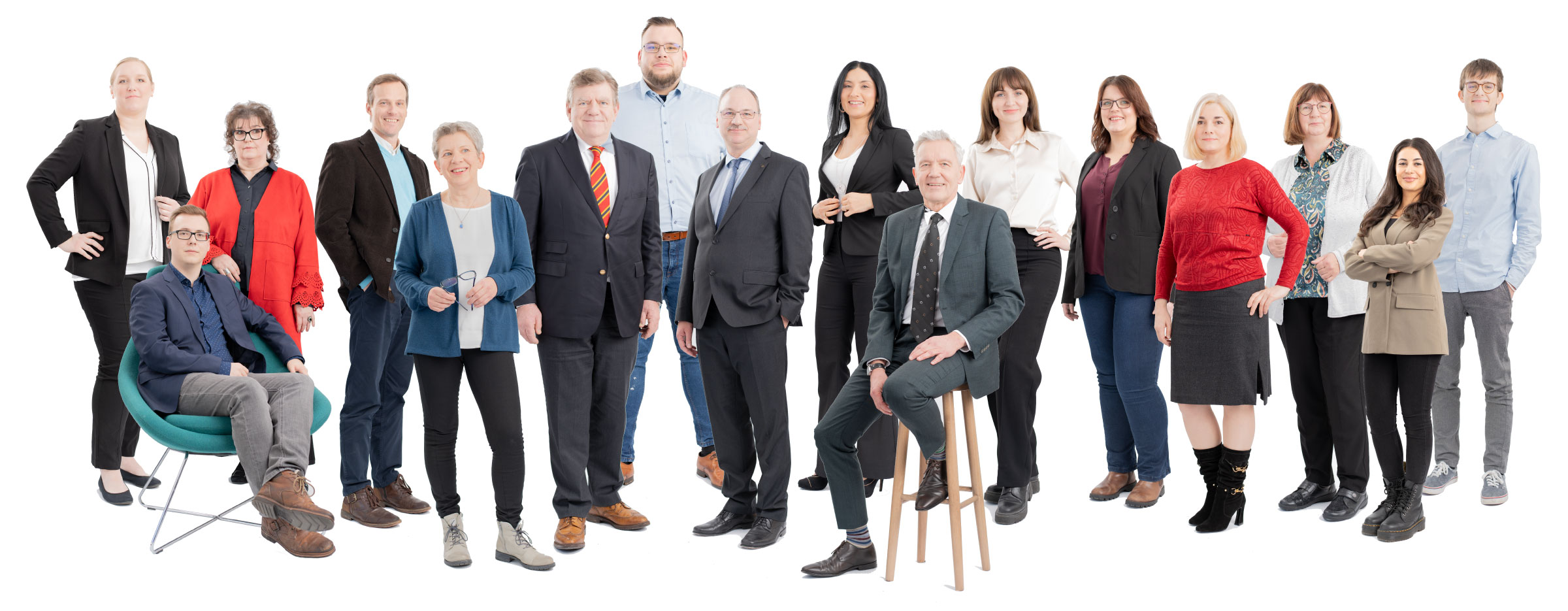 Das Bild zeigt eine größere Gruppe von Menschen unterschiedlichen Alters, die nebeneinander stehen und teilweise sitzen, vor einem weißen Hintergrund. Alle Personen tragen überwiegend Business- oder Business-Casual-Kleidung und blicken freundlich in die Kamera.